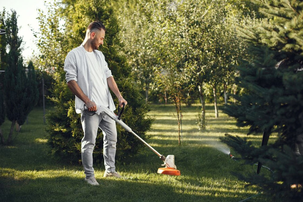 A man pushes a lawn mower across a grassy yard, with a clear blue sky and a fence visible in the background.