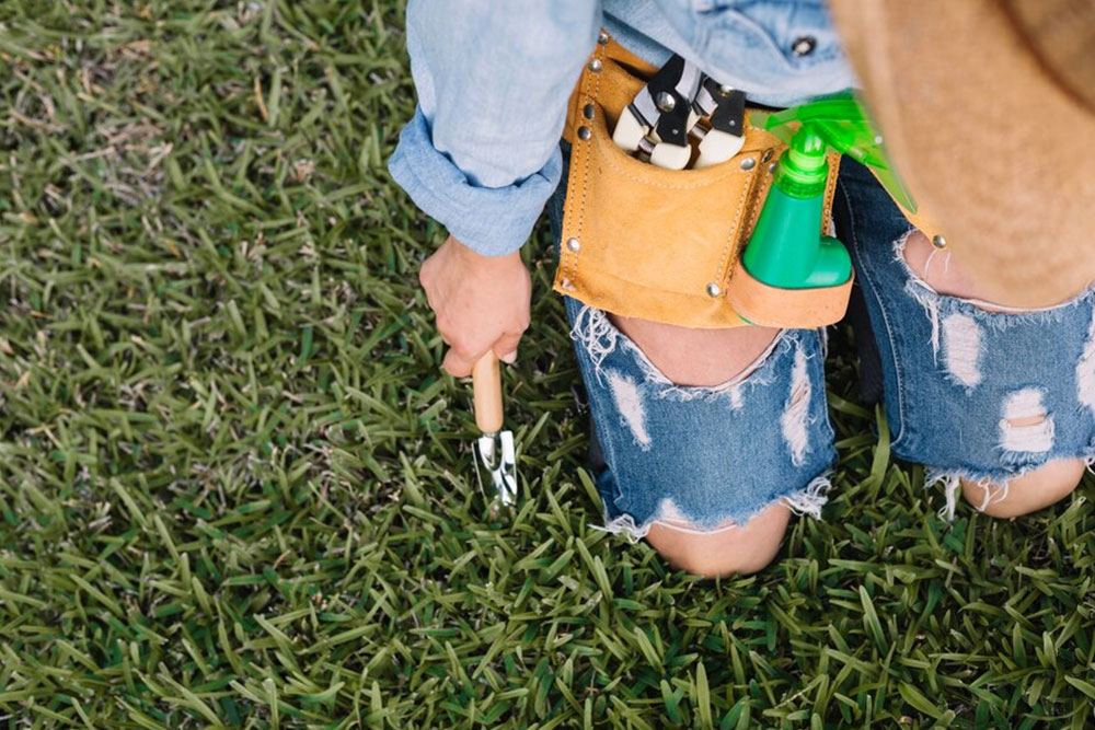 A woman wearing a hat and jeans kneels on the grass, holding a gardening tool in her hands.