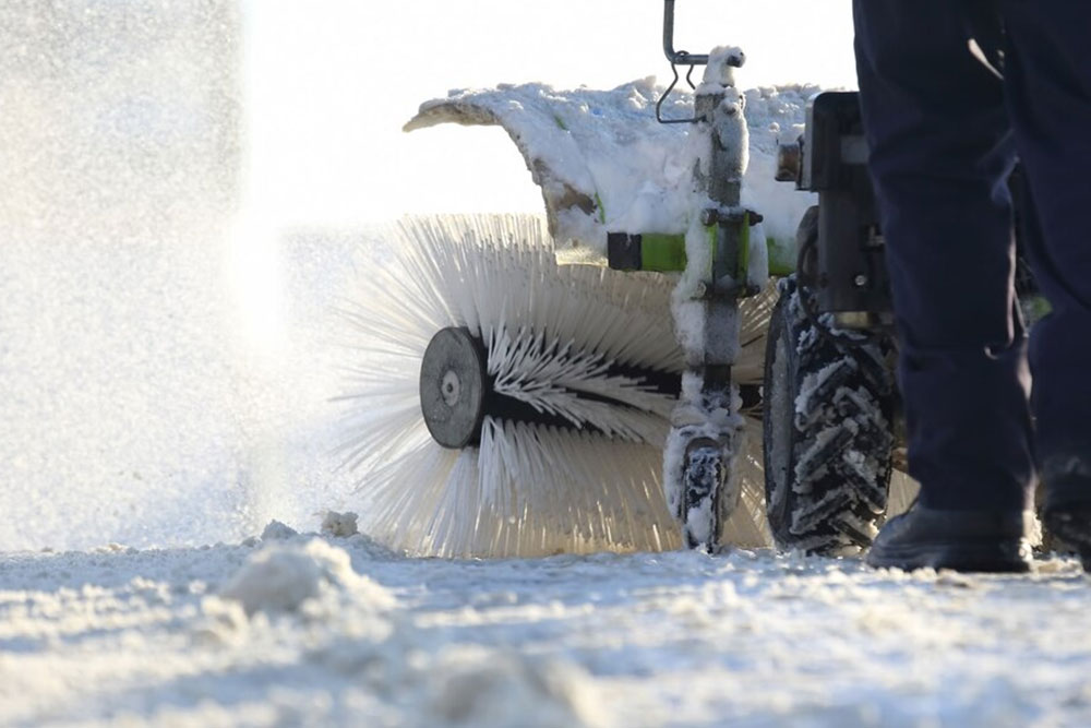 A man uses a snow blower to remove snow from a driveway, surrounded by a wintery scene.